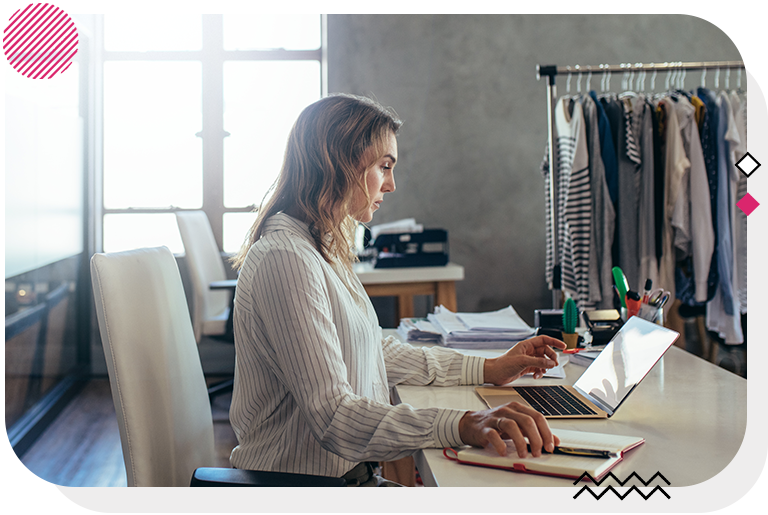 Woman sitting at a desk and looking at a laptop with a full clothes rack in the background